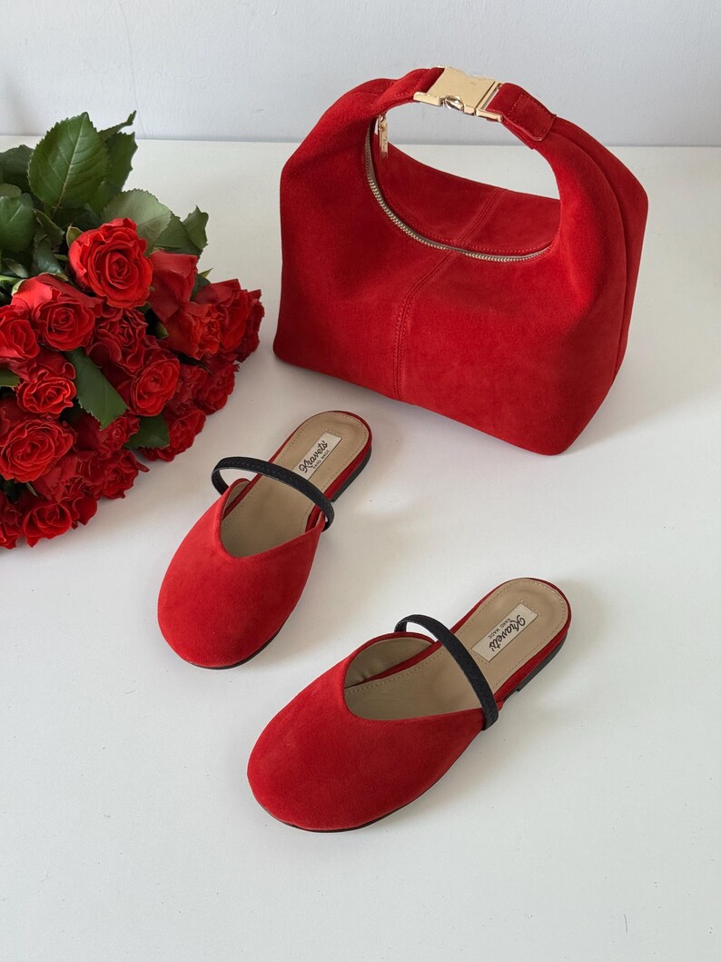 K&ouml;nnte beinhalten: Eine rote Wildlederhandtasche mit goldfarbener Schnalle und Rei&szlig;verschluss, kombiniert mit passenden roten Wildleder-Slippern mit schwarzen Riemen. Ein Strau&szlig; roter Rosen ist ebenfalls zu sehen.