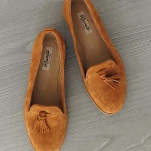 K&ouml;nnte beinhalten: Zwei orangefarbene Wildleder-Loafer mit Quasten. Die Schuhe haben eine runde Spitze und einen niedrigen Absatz. Der Markenname "Kravets" ist auf der Innensohle sichtbar. Pr&auml;sentiert auf einer hellgrauen Oberfl&auml;che.