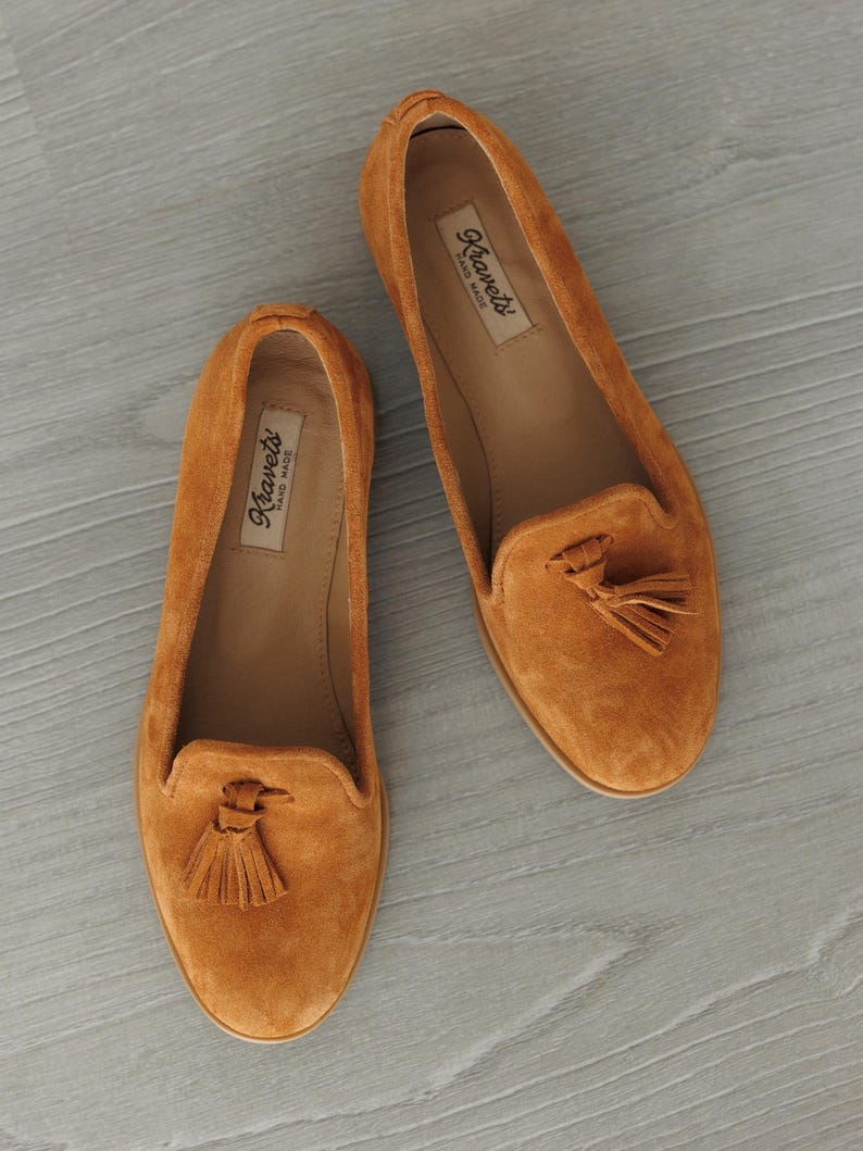 K&ouml;nnte beinhalten: Zwei orangefarbene Wildleder-Loafer mit Quasten. Die Schuhe haben eine runde Spitze und einen niedrigen Absatz. Der Markenname "Kravets" ist auf der Innensohle sichtbar. Pr&auml;sentiert auf einer hellgrauen Oberfl&auml;che.