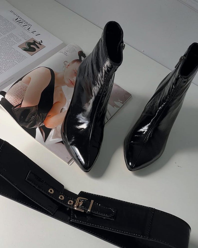 May include: A pair of glossy black ankle boots with pointed toes and a zipper closure. A black belt with a gold-colored buckle is also visible. The boots are placed on a white surface next to a magazine.
