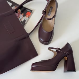 May include: Burgundy leather Mary Jane platform heels with a buckle closure. A matching burgundy tote bag with a handle and a magazine are also in the image. The heels have a chunky block heel.
