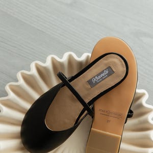 K&ouml;nnte beinhalten: Schwarze Lederballerinas mit Riemen, die auf einer wei&szlig;en, gewellten Schale liegen. Die Sohle ist beige mit dem Text "ITALY DESIGN" und der Gr&ouml;&szlig;e 37. Die Ballerinas tragen das Etikett "Kravets HAND MADE".