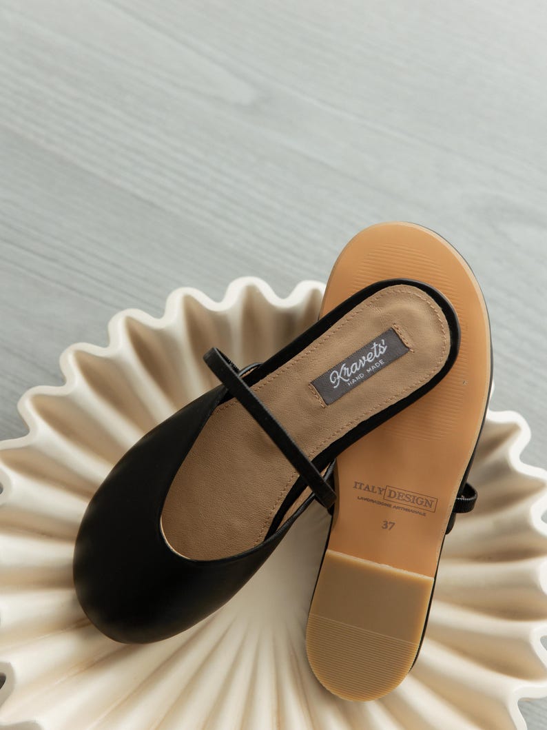 K&ouml;nnte beinhalten: Schwarze Lederballerinas mit Riemen, die auf einer wei&szlig;en, gewellten Schale liegen. Die Sohle ist beige mit dem Text "ITALY DESIGN" und der Gr&ouml;&szlig;e 37. Die Ballerinas tragen das Etikett "Kravets HAND MADE".
