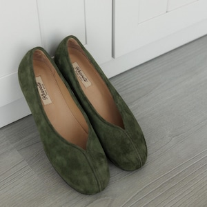 May include: A pair of olive green suede ballet flats. The shoes have a rounded toe and a low-cut vamp. The interior of the shoe is a light beige colour. The brand name "Klavets" is printed on the insole. The shoes are placed on a light grey wooden floor.