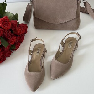 May include: A taupe suede crossbody bag with a flap closure, a pair of matching slingback heels, and a bouquet of red roses. The shoes have pointed toes and a small heel. The bag has a silver chain strap.