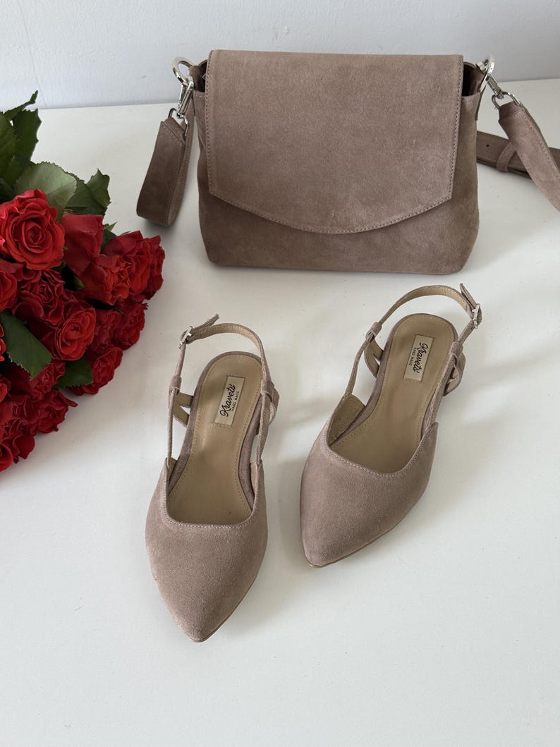 May include: A taupe suede crossbody bag with a flap closure, a pair of matching slingback heels, and a bouquet of red roses. The shoes have pointed toes and a small heel. The bag has a silver chain strap.