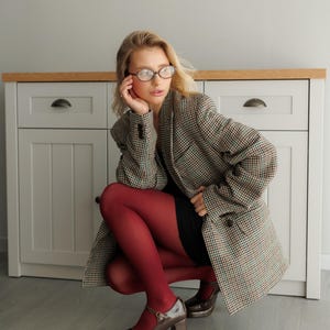 May include: A woman wearing a brown and white plaid blazer, a black dress, and red tights. She is wearing brown heeled shoes with a strap across the ankle. She is sitting on the floor in front of a white cabinet with a wooden top.