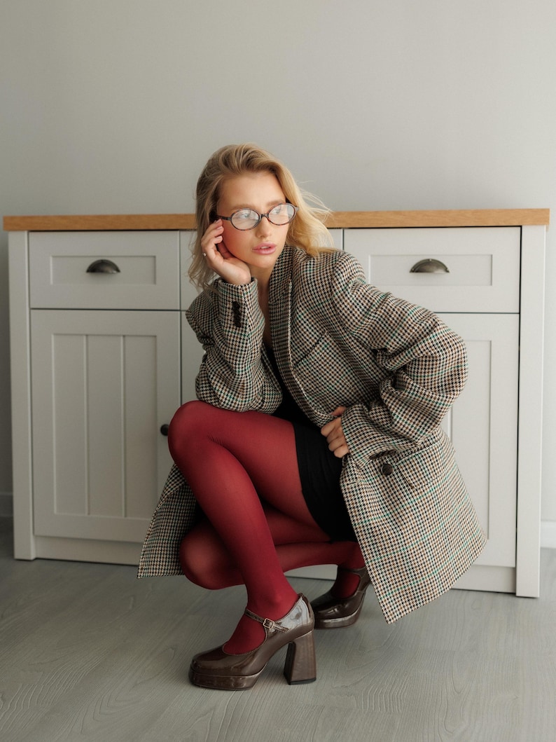 May include: A woman wearing a brown and white plaid blazer, a black dress, and red tights. She is wearing brown heeled shoes with a strap across the ankle. She is sitting on the floor in front of a white cabinet with a wooden top.