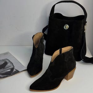 May include: A black suede handbag with a top handle and a crossbody strap, next to a pair of black suede ankle boots with a pointed toe and a wooden heel. The boots have a zipper on the side. A magazine is on the left.