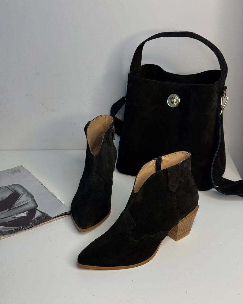 May include: A black suede handbag with a top handle and a crossbody strap, next to a pair of black suede ankle boots with a pointed toe and a wooden heel. The boots have a zipper on the side. A magazine is on the left.