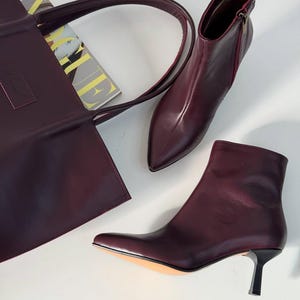 May include: A burgundy leather tote bag with a long handle, a pair of burgundy leather ankle boots with pointed toes and a small heel, and a magazine. The boots have a zipper on the side.