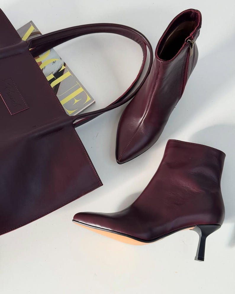 May include: A burgundy leather tote bag with a long handle, a pair of burgundy leather ankle boots with pointed toes and a small heel, and a magazine. The boots have a zipper on the side.