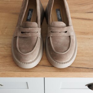 May include: A pair of light brown suede loafers with a thick platform sole. The loafers have a leather strap across the top and the brand name "Rieker" is printed on the inside of the shoe.
