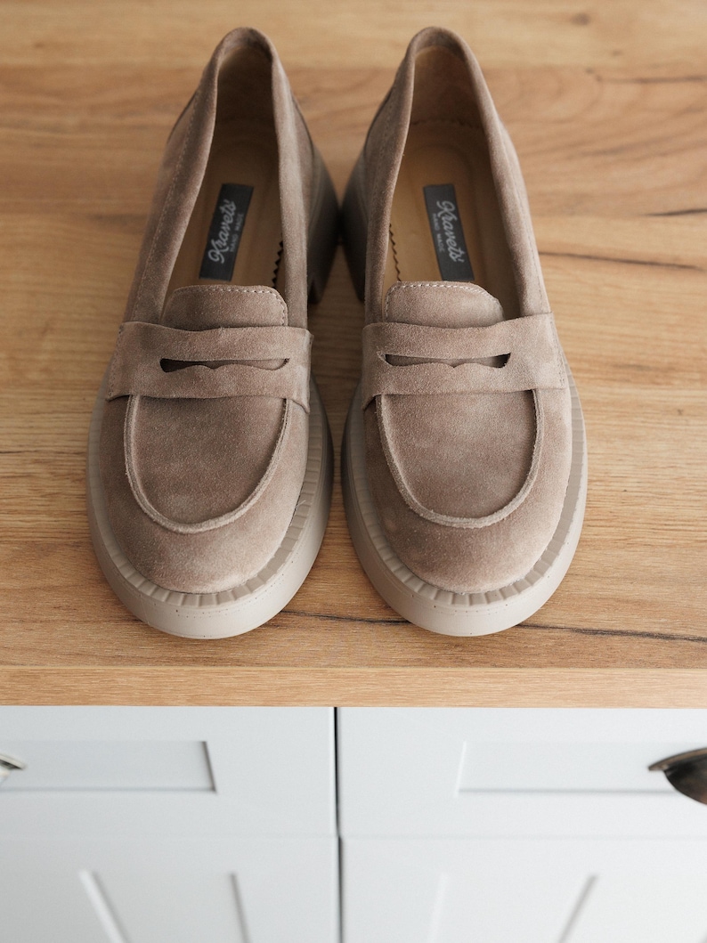 May include: A pair of light brown suede loafers with a thick platform sole. The loafers have a leather strap across the top and the brand name "Rieker" is printed on the inside of the shoe.