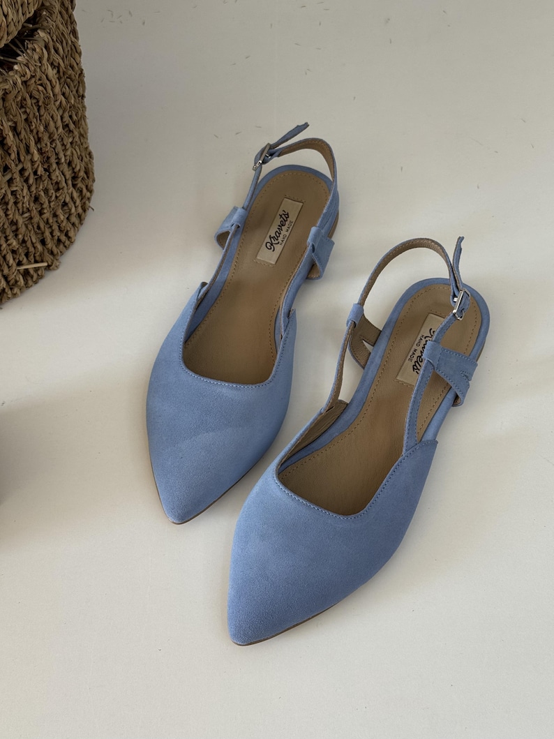 May include: A pair of light blue suede flats with pointed toes and slingback straps. The shoes have a small buckle closure and a neutral-toned insole. The brand name "Khavels" is visible on the insole. The shoes are displayed on a white surface.