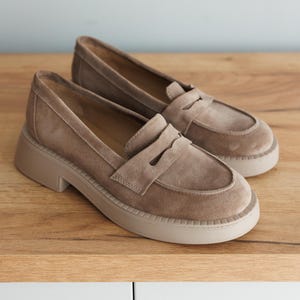 May include: A pair of light brown suede loafers with a chunky platform sole. The loafers have a classic penny loafer design with a strap across the top.