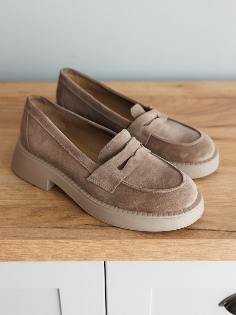 May include: A pair of light brown suede loafers with a chunky platform sole. The loafers have a classic penny loafer design with a strap across the top.