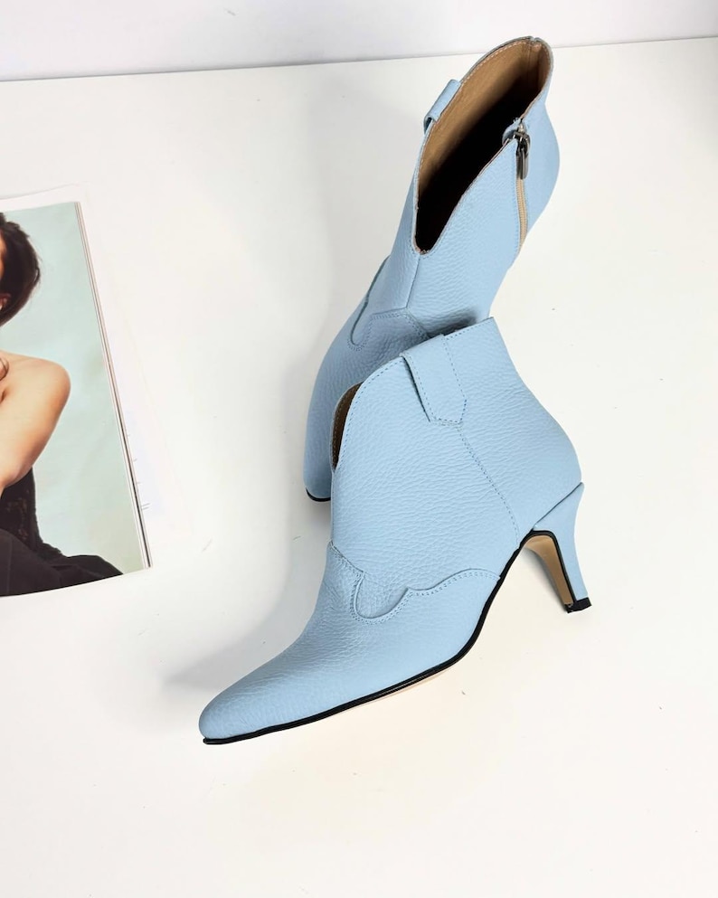 May include: A pair of light blue ankle boots with a pointed toe and a small heel. The boots have a western-inspired design with decorative stitching and a side zipper. The heel measures approximately 2.5 inches.