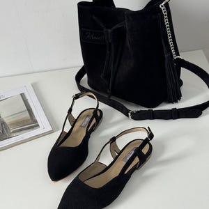 May include: A black suede bucket bag with a drawstring closure and a shoulder strap, paired with matching black slingback flats. The flats have pointed toes and small heels. The bag has a silver chain detail and a tassel.