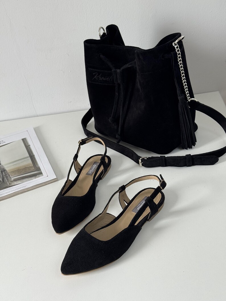 May include: A black suede bucket bag with a drawstring closure and a shoulder strap, paired with matching black slingback flats. The flats have pointed toes and small heels. The bag has a silver chain detail and a tassel.