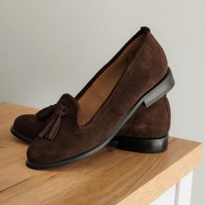 K&ouml;nnte beinhalten: Ein Paar braune Wildleder-Loafer mit Quasten. Die Schuhe haben einen niedrigen Absatz und ein klassisches Design. Die Loafer werden auf einer Holzoberfl&auml;che pr&auml;sentiert, die ihre satte Farbe und Textur hervorhebt. Eine stilvolle Schuhoption.