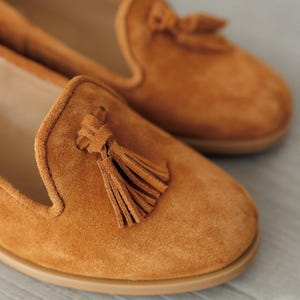 K&ouml;nnte beinhalten: Ein Paar Wildleder-Loafer in einem warmen, goldbraunen Farbton. Jeder Schuh hat eine dekorative Quaste und eine abgerundete Spitze. Die Sohlen sind hellbraun. Die Schuhe scheinen ungetragen zu sein.