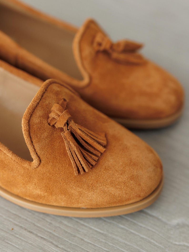 K&ouml;nnte beinhalten: Ein Paar Wildleder-Loafer in einem warmen, goldbraunen Farbton. Jeder Schuh hat eine dekorative Quaste und eine abgerundete Spitze. Die Sohlen sind hellbraun. Die Schuhe scheinen ungetragen zu sein.