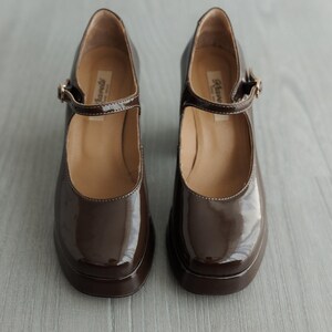 May include: A pair of brown patent leather shoes with a buckle strap. The shoes have a chunky heel and a rounded toe.