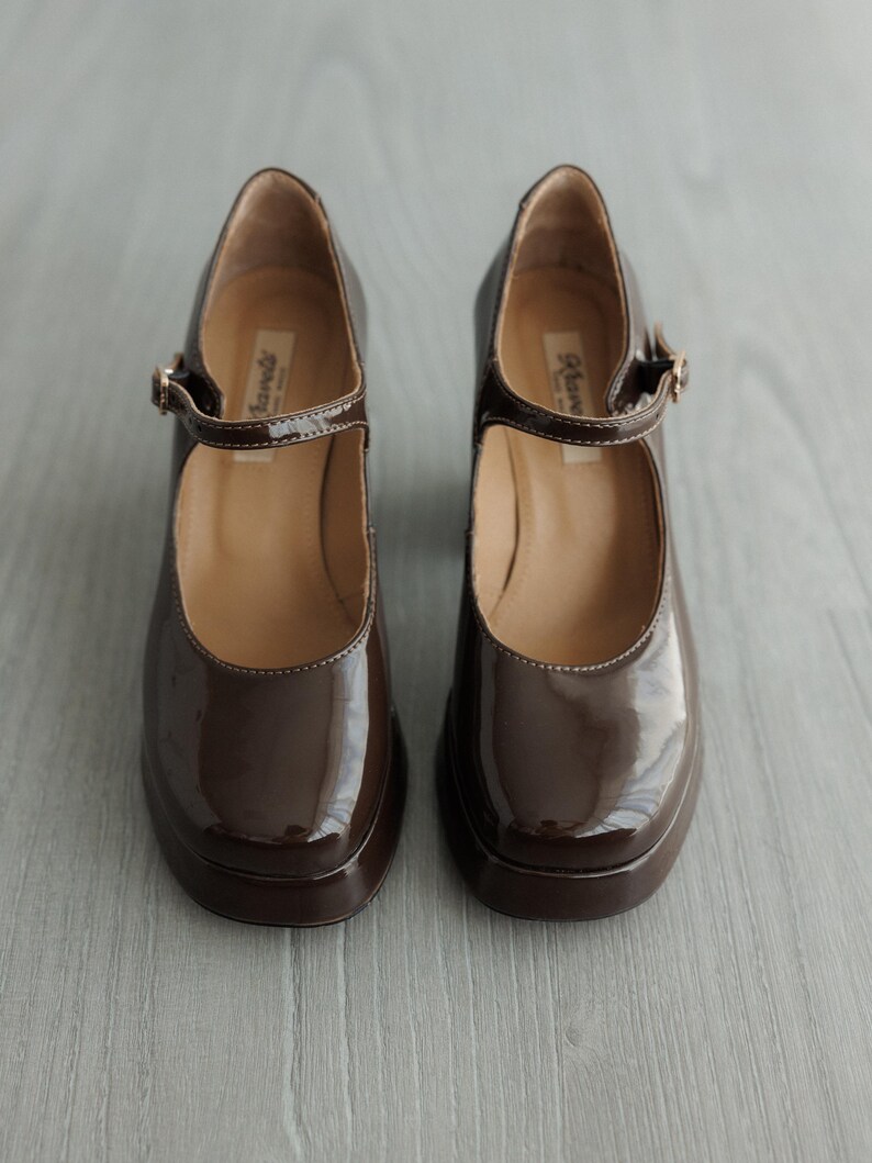 May include: A pair of brown patent leather shoes with a buckle strap. The shoes have a chunky heel and a rounded toe.