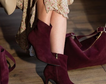 May include: Burgundy suede ankle boots with a pointed toe and a small heel. The boots have a side zipper and are paired with a matching handbag. The image shows a floral dress and a tan coat.