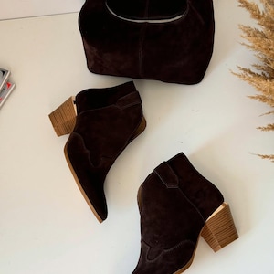 May include: A dark brown suede handbag with a gold-colored clasp and zipper, paired with matching ankle boots. The boots have a pointed toe and a wooden block heel. The ensemble is set against a white background, showcasing a coordinated fashion statement.
