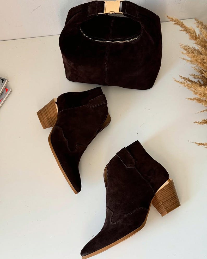 May include: A dark brown suede handbag with a gold-colored clasp and zipper, paired with matching ankle boots. The boots have a pointed toe and a wooden block heel. The ensemble is set against a white background, showcasing a coordinated fashion statement.