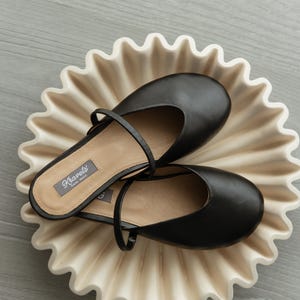K&ouml;nnte beinhalten: Ein Paar schwarze Leder-Ballerinas mit Riemen auf einer cremefarbenen Keramikplatte mit gewelltem Rand. Die Schuhe haben eine runde Spitze und einen kleinen Absatz. Die Platte befindet sich auf einer hellgrauen Oberfl&auml;che.