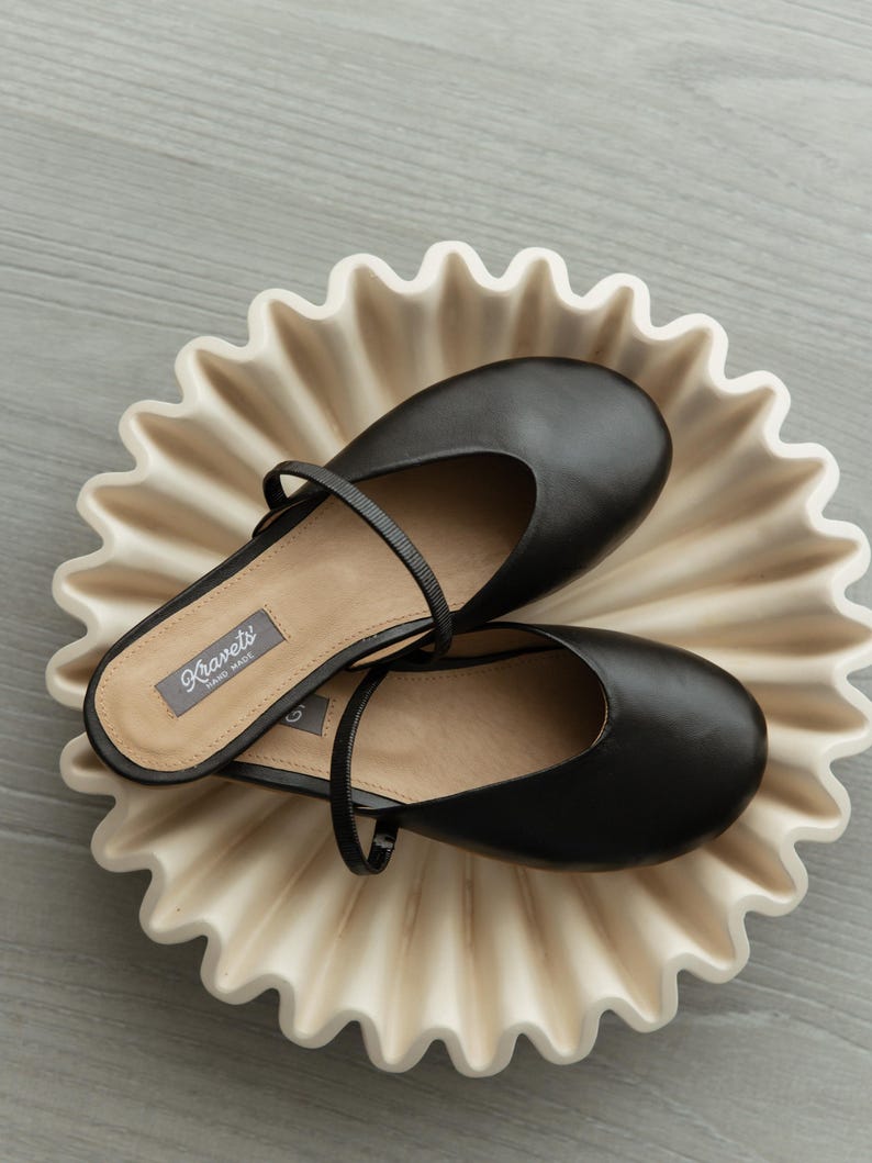 K&ouml;nnte beinhalten: Ein Paar schwarze Leder-Ballerinas mit Riemen auf einer cremefarbenen Keramikplatte mit gewelltem Rand. Die Schuhe haben eine runde Spitze und einen kleinen Absatz. Die Platte befindet sich auf einer hellgrauen Oberfl&auml;che.