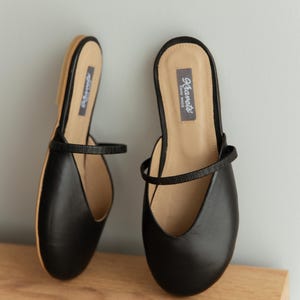 K&ouml;nnte beinhalten: Ein Paar schwarze Leder-Ballerinas mit einem Riemen &uuml;ber dem Oberteil. Die Schuhe haben eine runde Spitze und eine beige Innensohle. Der Markenname "Kiarets" ist auf der Innensohle aufgedruckt. Die Schuhe werden auf einer Holzoberfl&auml;che pr&auml;sentiert.
