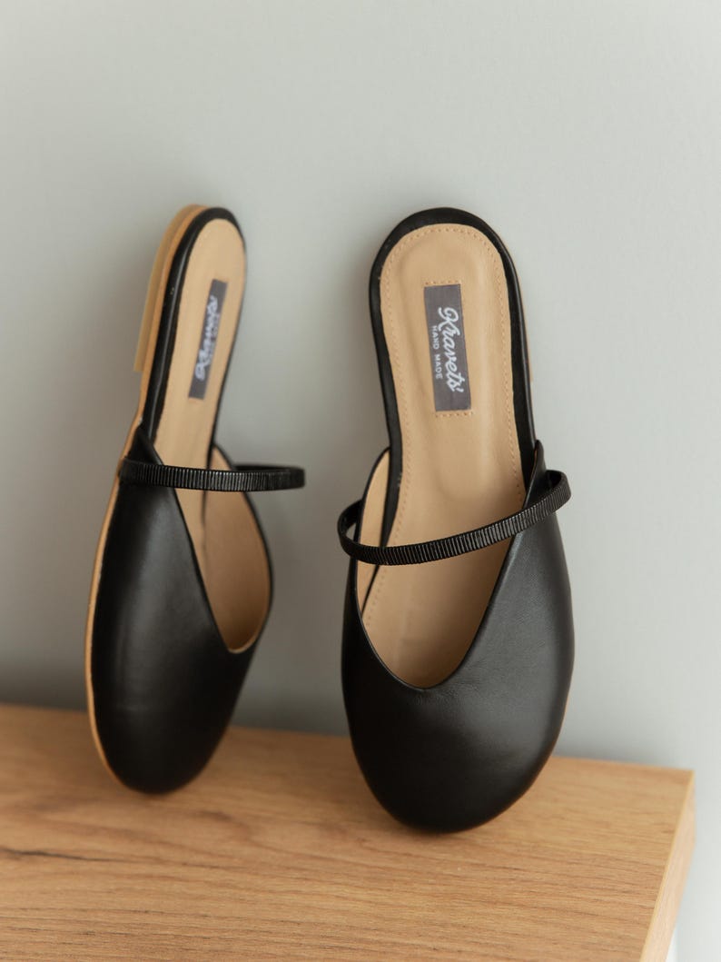 K&ouml;nnte beinhalten: Ein Paar schwarze Leder-Ballerinas mit einem Riemen &uuml;ber dem Oberteil. Die Schuhe haben eine runde Spitze und eine beige Innensohle. Der Markenname "Kiarets" ist auf der Innensohle aufgedruckt. Die Schuhe werden auf einer Holzoberfl&auml;che pr&auml;sentiert.