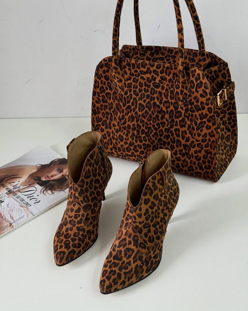May include: A pair of leopard print ankle boots with pointed toes and high heels, next to a matching handbag. The bag has a rectangular shape with handles and a gold buckle. A magazine is in the background.