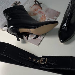 May include: Black patent leather ankle boots with pointed toes and stiletto heels. A matching black patent leather wide belt with a silver buckle is also visible. The items are displayed on a white surface with a magazine in the background.