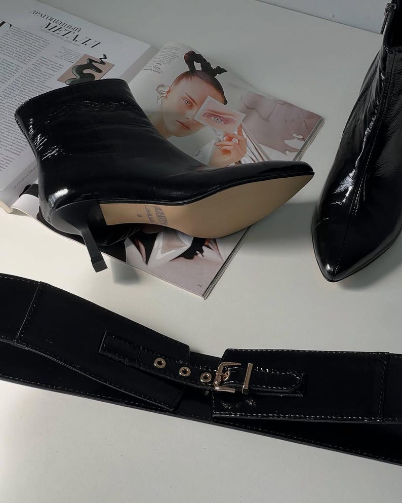 May include: Black patent leather ankle boots with pointed toes and stiletto heels. A matching black patent leather wide belt with a silver buckle is also visible. The items are displayed on a white surface with a magazine in the background.