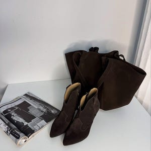 May include: A pair of dark brown suede ankle boots with pointed toes and a small heel, next to a matching tote bag. A magazine with black and white images is on the left.