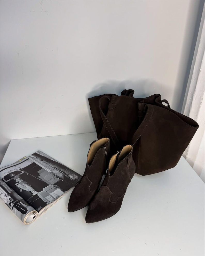 May include: A pair of dark brown suede ankle boots with pointed toes and a small heel, next to a matching tote bag. A magazine with black and white images is on the left.