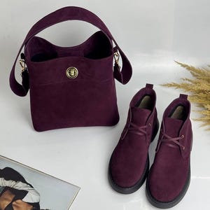 Puede incluir: Un conjunto a juego de accesorios de ante burdeos, que incluye un bolso con cierre dorado y un par de botines con suelas negras. El bolso tiene un asa superior y una correa de hombro desmontable. Las botas tienen un diseño con cordones.