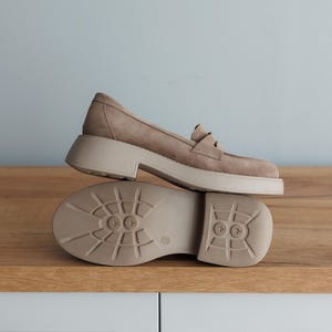 May include: A pair of light brown suede loafers with a chunky platform sole. The loafers have a classic loafer design with a buckle detail on the top.