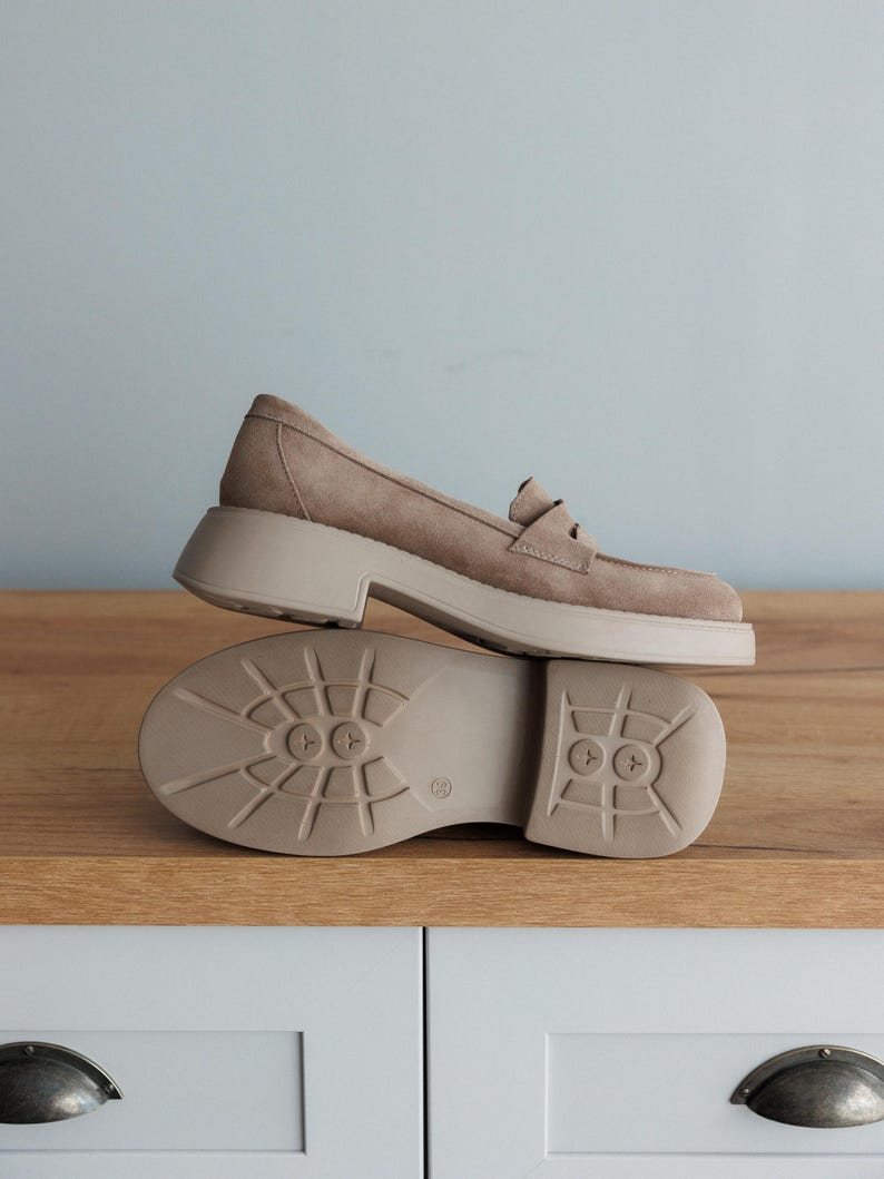 May include: A pair of light brown suede loafers with a chunky platform sole. The loafers have a classic loafer design with a buckle detail on the top.