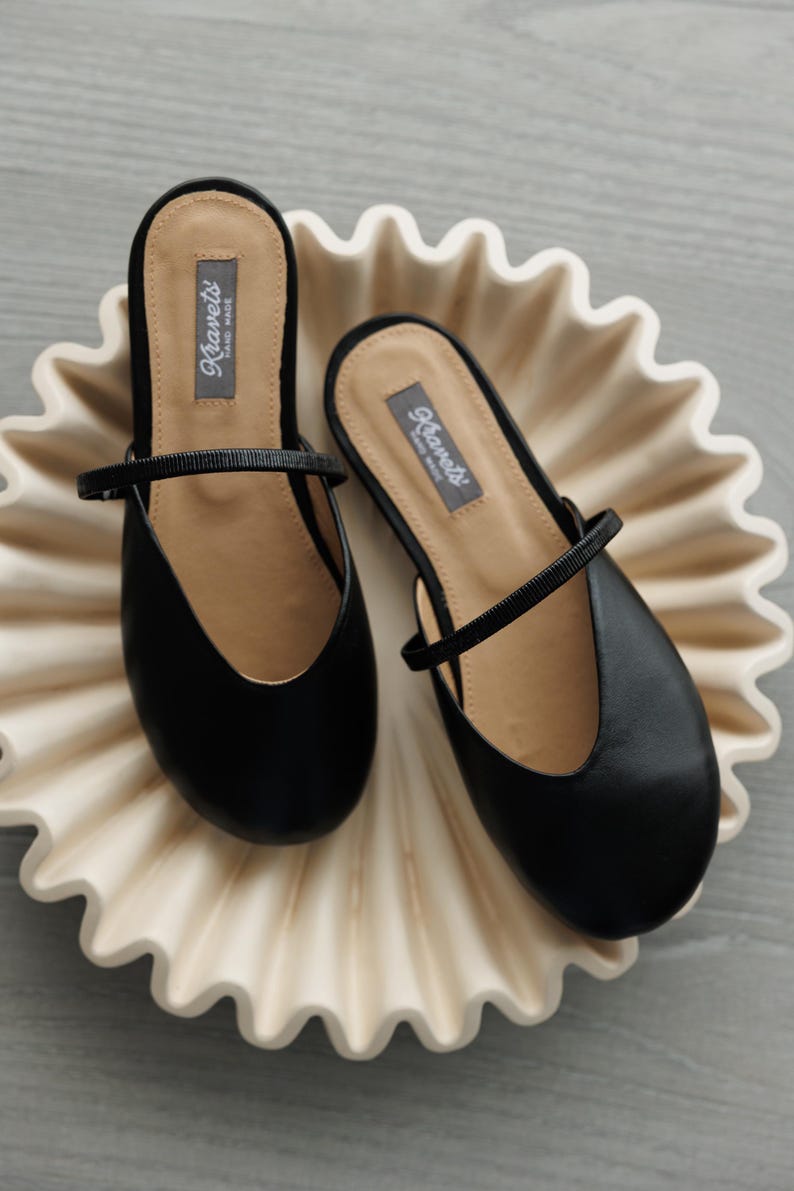 K&ouml;nnte beinhalten: Ein Paar schwarze Slipper mit Riemen, pr&auml;sentiert auf einer dekorativen, cremefarbenen, wellenf&ouml;rmigen Schale. Die Schuhe haben eine runde Spitze und einen kleinen Absatz. Der Markenname "Ravel's" ist auf der Innensohle sichtbar.
