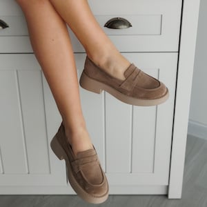 May include: A pair of brown suede loafers with a chunky platform sole. The loafers have a simple design with a single strap across the vamp.