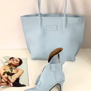 May include: A light blue leather tote bag and matching ankle boots with a pointed toe and a small heel. The tote bag has a rectangular label with the word "Kismet's" embossed on it. A magazine with a woman holding a rose is in the background.