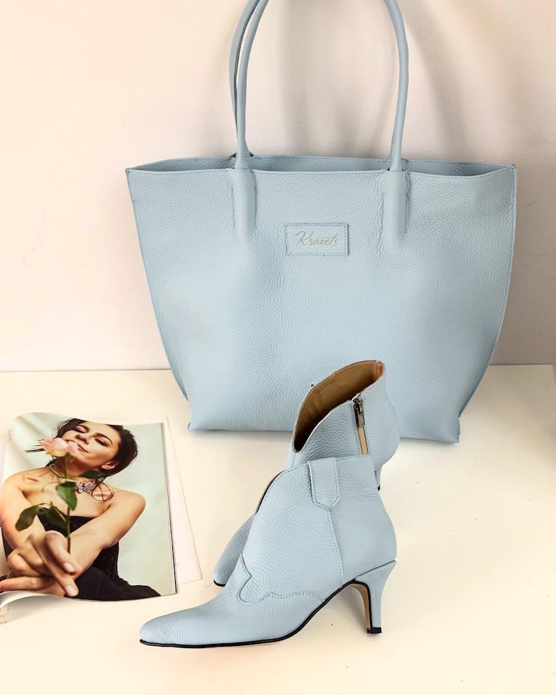 May include: A light blue leather tote bag and matching ankle boots with a pointed toe and a small heel. The tote bag has a rectangular label with the word "Kismet's" embossed on it. A magazine with a woman holding a rose is in the background.