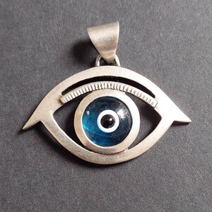 Könnte beinhalten: Ein silberner Anhänger in Form eines Auges mit einem blauen und weißen bösen Blick-Anhänger. Der Anhänger ist ein beliebtes Symbol für Schutz vor Unglück.