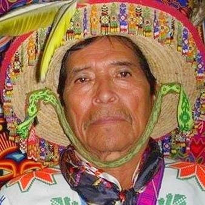 Pode incluir: Um homem usando um chap&eacute;u de palha com acabamentos decorativos coloridos e uma tira de queixo verde. O chap&eacute;u apresenta uma variedade de padr&otilde;es e desenhos, incluindo o que parecem ser penas. O homem tamb&eacute;m est&aacute; usando uma camisa estampada.